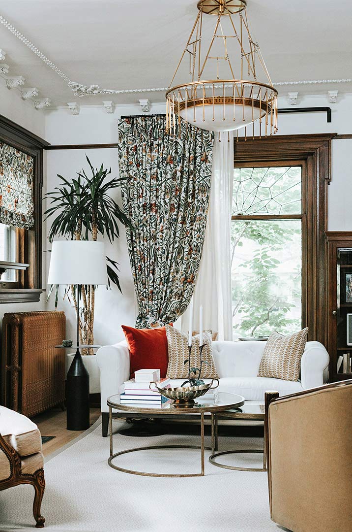Patterned curtains frame a traditional window and white sofa with velvet red pillow behind a brass finished nesting tables. 