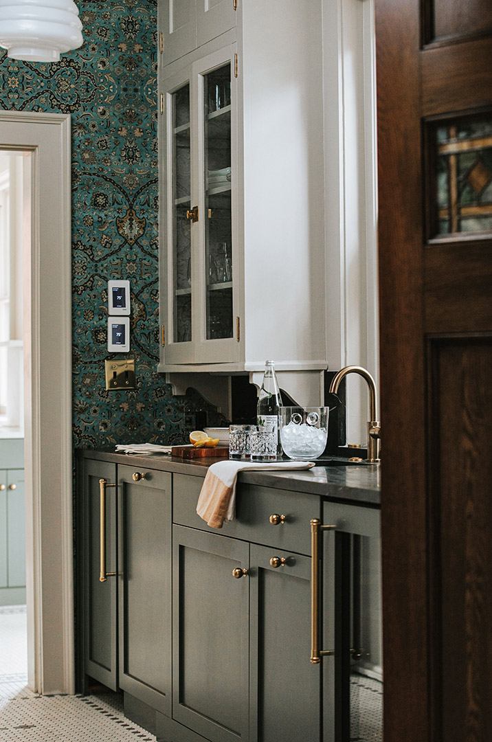 Green cabinets provide a base for soapstone countertops, brass fixtures, and rich teal colored wallpaper. 