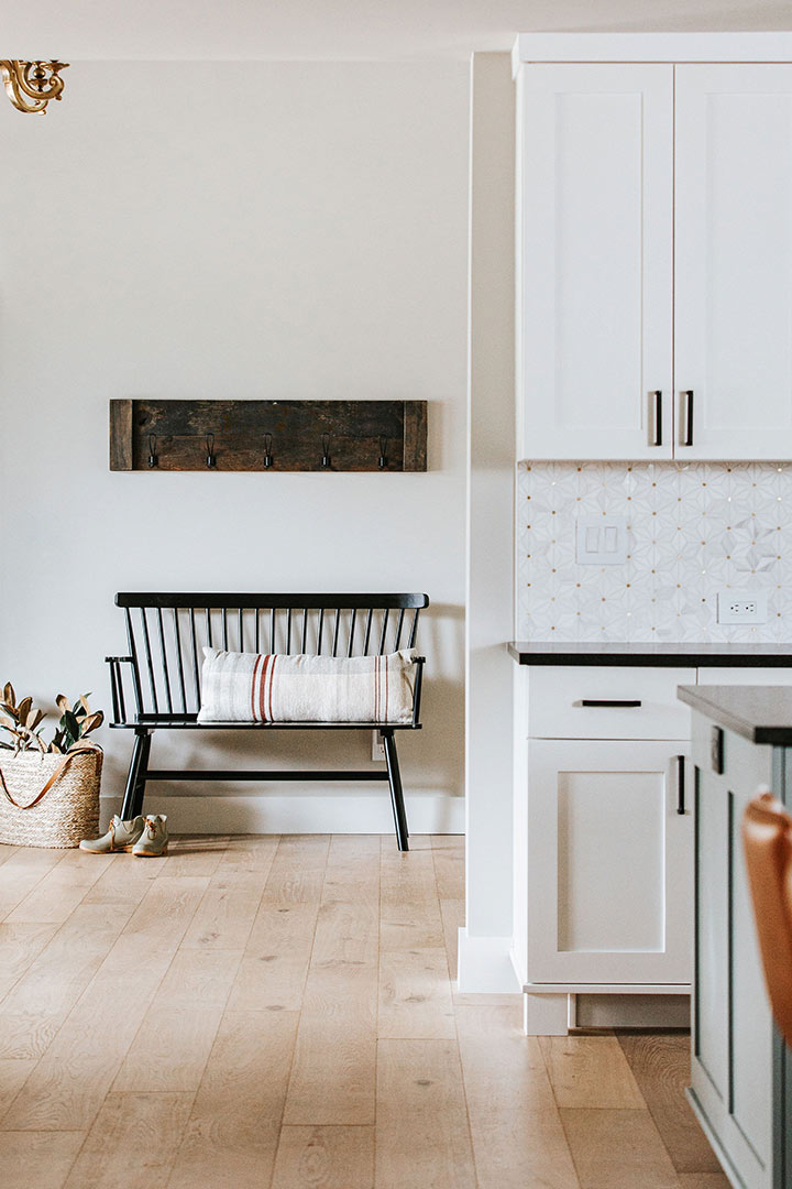 Bright entry way with a farmhouse bench shows a glimpse into the kitchen and it's white shaker cabinets