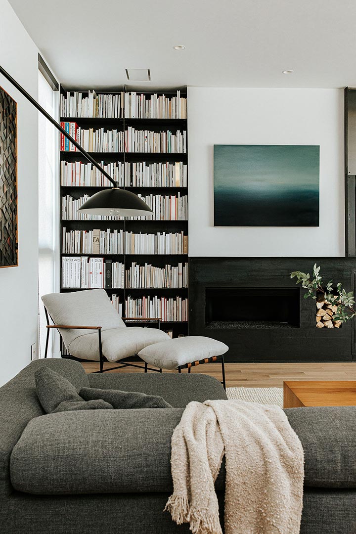 A modern floor to ceiling bookcase and window frame the dark black rolled steele fireplace, paired in this mindful interior design by J. Reiko Design + Co
