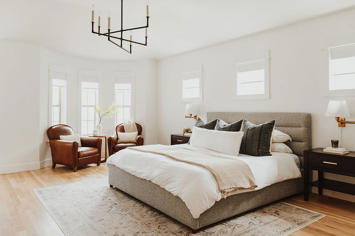 A modern classic bedroom with leather chairs, an aged iron chandelier and neutral gray and white bedding