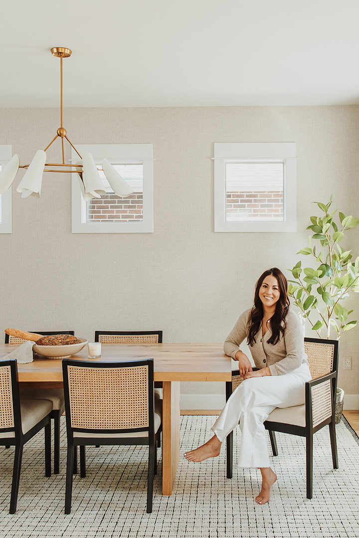 Designer Jenny Murphy sits at a white oak dining table in caned back chairs she chose for this modern minimalist whole house remodel in Denver, Colorado