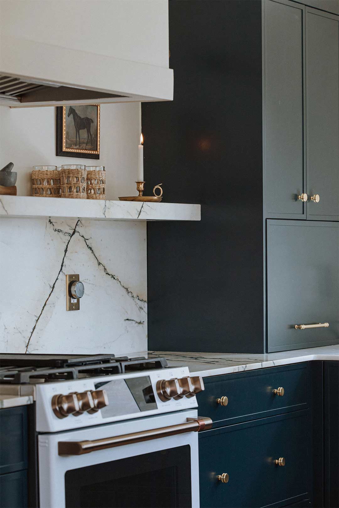 Dark blue cabinets with brass pulls, marble countertops and backsplash with a marble ledge on a range wall.