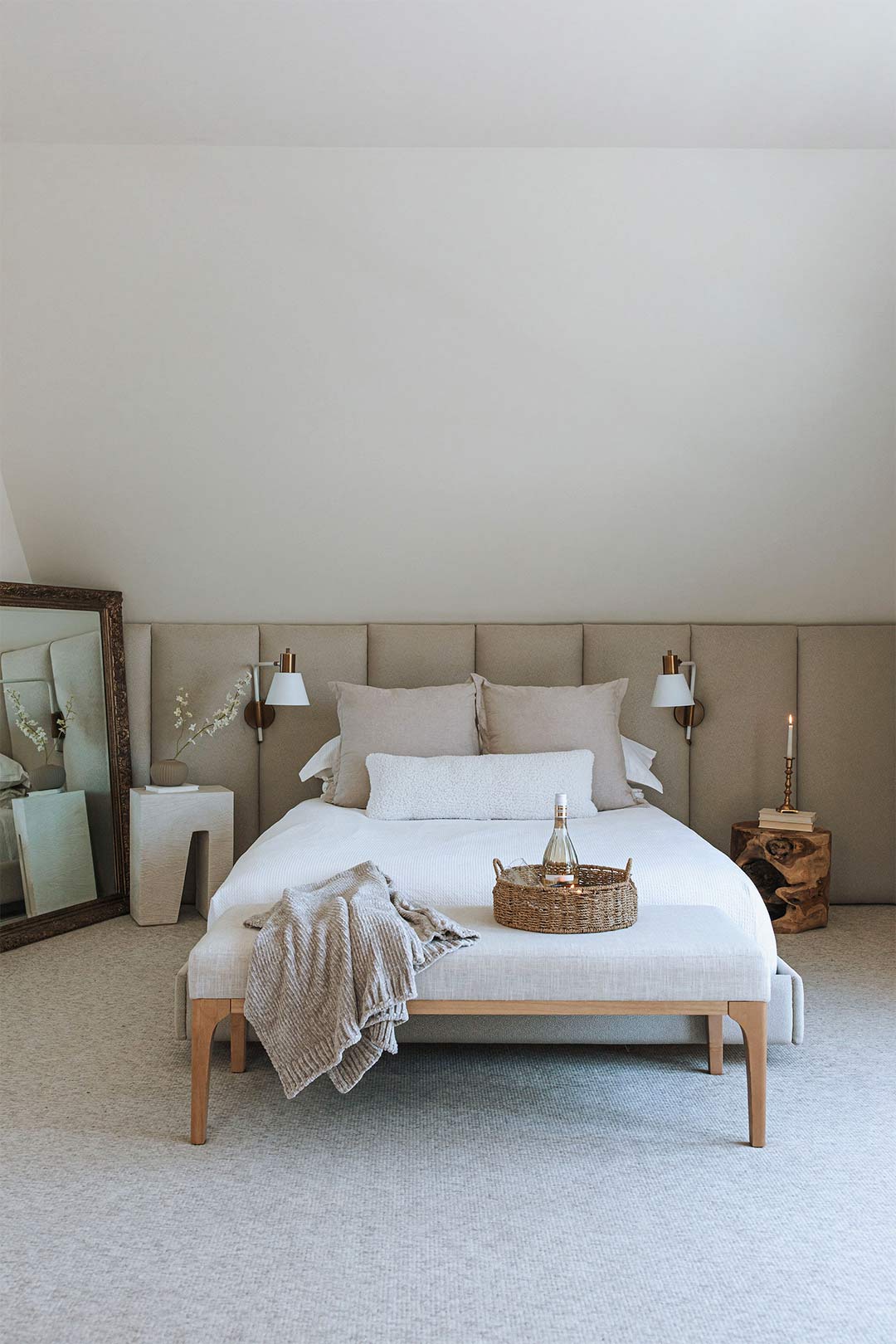 Full-wall upholstered heardboard in a neutral toned bedroom with modern brass scones, and organic-modern furniture.