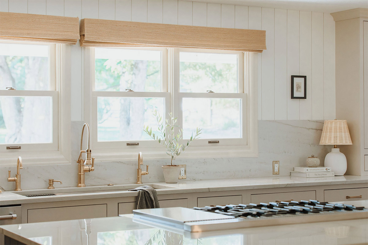 Woven Roman Shades span the windows of a bright and airy modern farmhouse kitchen with light colored Quartzite Countertops with Slab Backsplash, brass hardware, and white paneling
