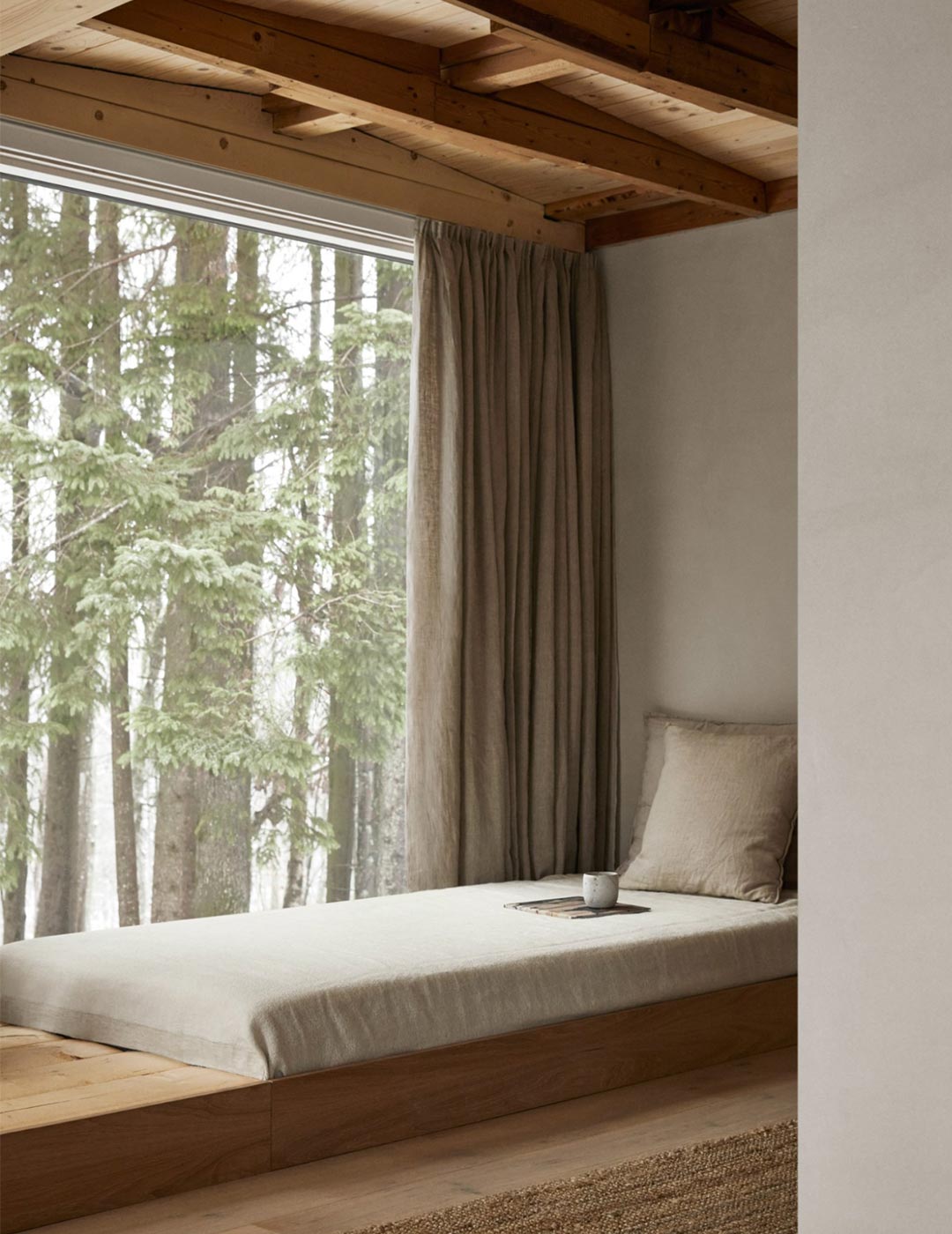 A cozy nook in a cabin designed by Norm Architects that shows a daybed with a large oversized floor to ceiling window with a view of a Forest.