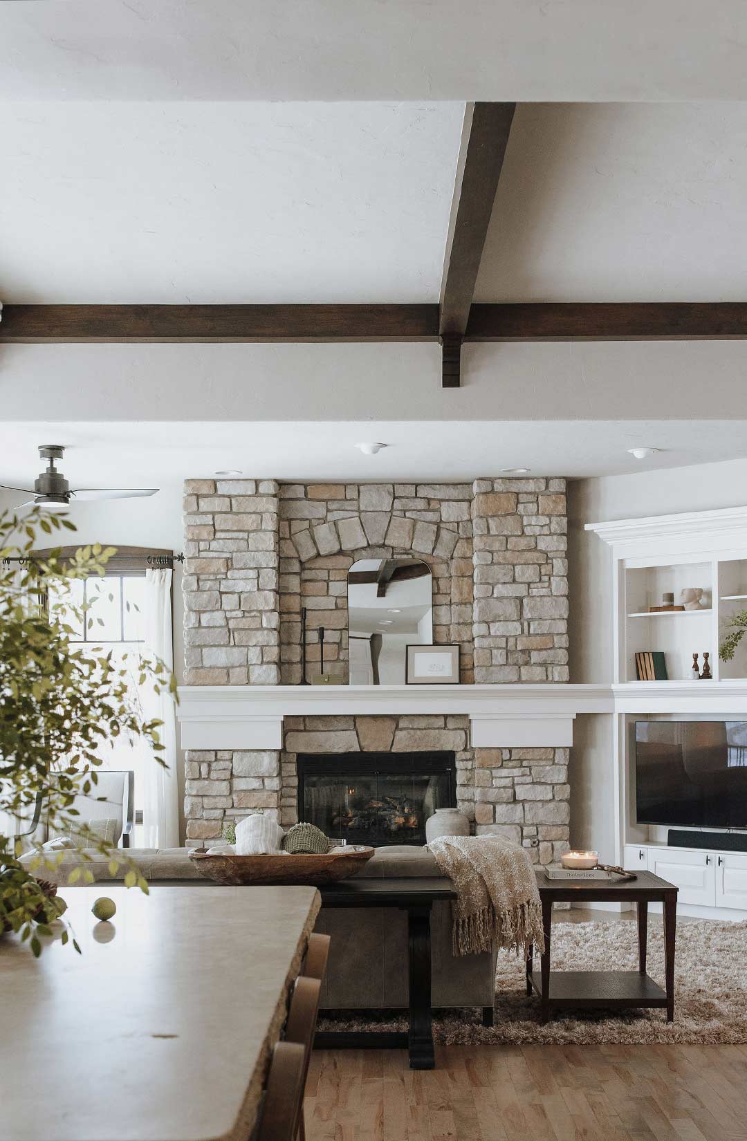 A straight on view of the floor to ceiling stone fireplace in a traditional living room that features dark wood ceiling beams, arched mirror on the mantel and the Petrel LED ceiling fan from Rejuvenation