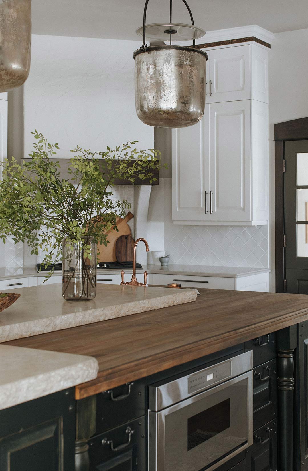 Visual Comfort Lorford Smoke Bell Lantern hangs over the Mediterranean island with a Travertine Countertop with Chiseled Edge that rests on top of a Butcher Block Countertop with a dark green island cabinet color