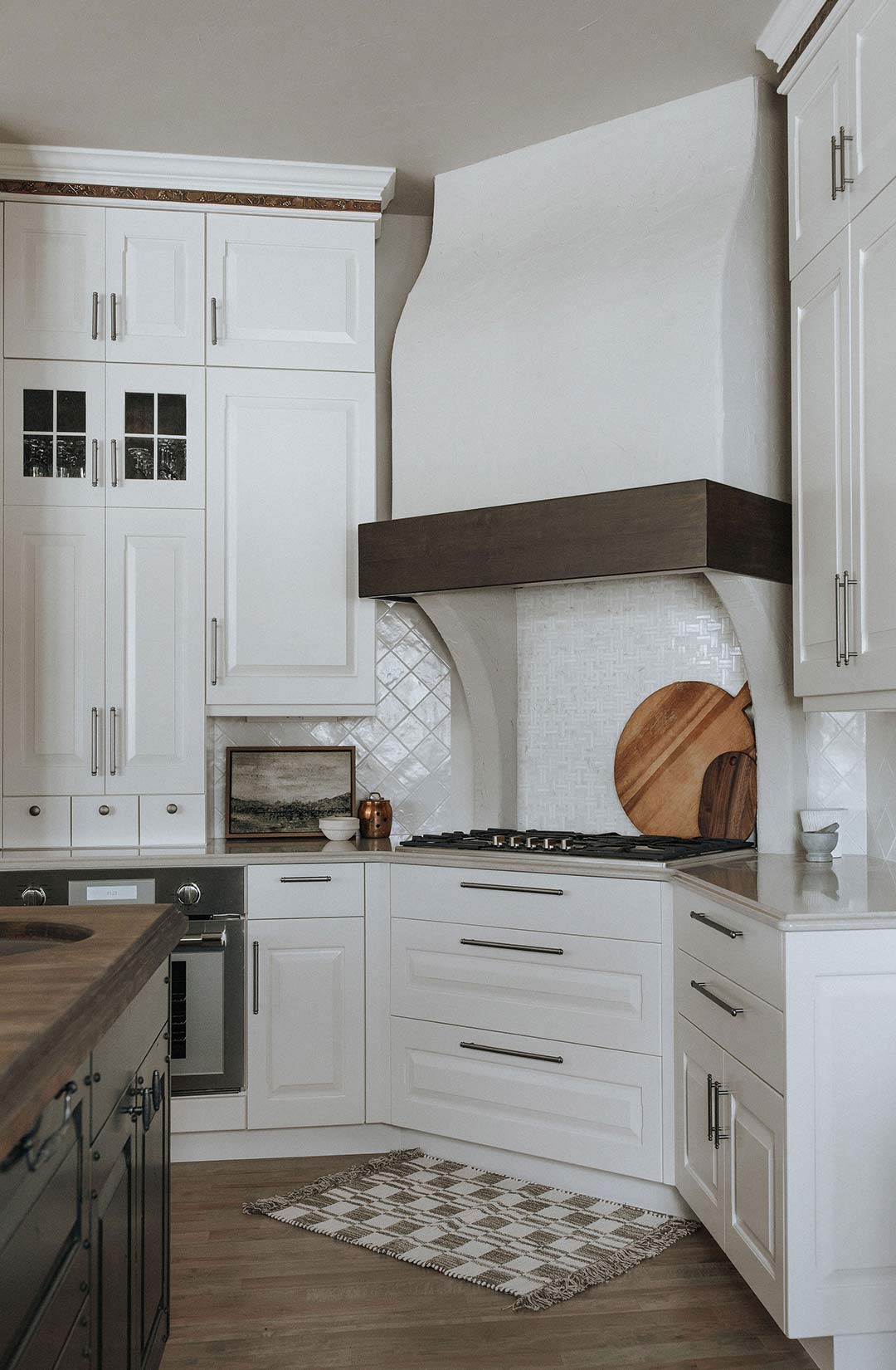 A curved Plaster hood vent with a dark wood trim is brightened up by the Zellige Tile Backsplash in Marazzi with off-white cabinets in Sherwin Williams SW6070 Heron Plume
