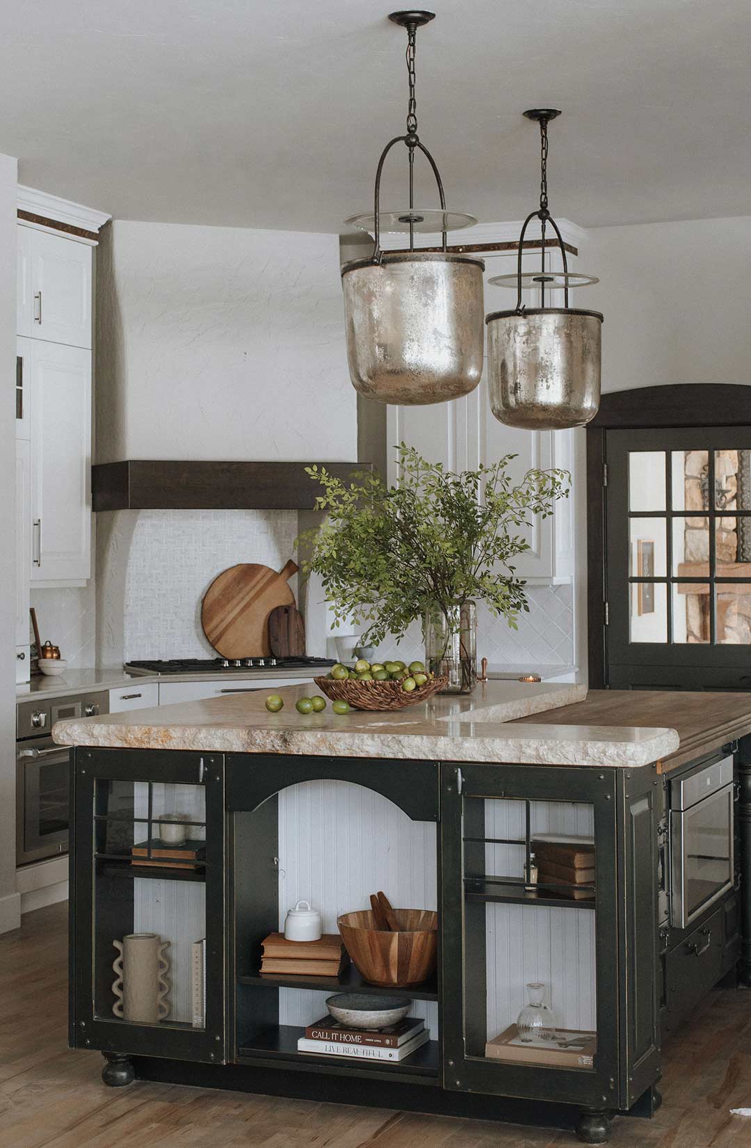 A large oversized island in a Mountain Mediterranean Kitchen features a unique, custom built storage unit and the end of the island with a Travertine Countertop with a Chiseled Edge and two Visual Comfort Lorford Smoke Bell Lantern pendant lights