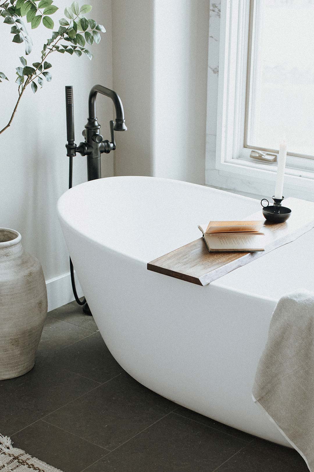 A free standing tub with a traditional floor mounted tub filler in matte black with a wood bath caddy with a book and a handheld candlestick carrier nodes to the historic charm and contemporary feeling of this primary bath remodel