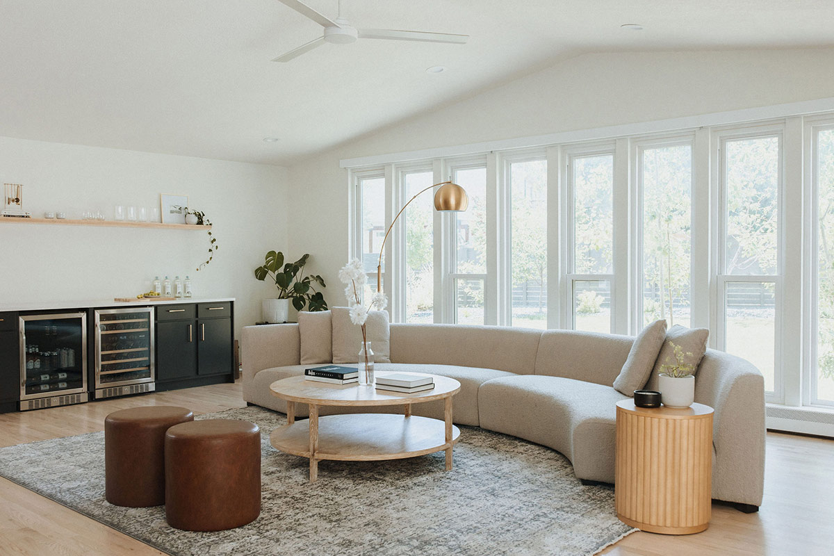 A modern living room features a Curved Sofa - Liam Sectional, a round white oak coffee table, and two brown leather ottomans. A brass dome floor lamp arches over the sofa. Large windows line the back wall, allowing natural light to fill the space. A bar area with black cabinets is on the left.