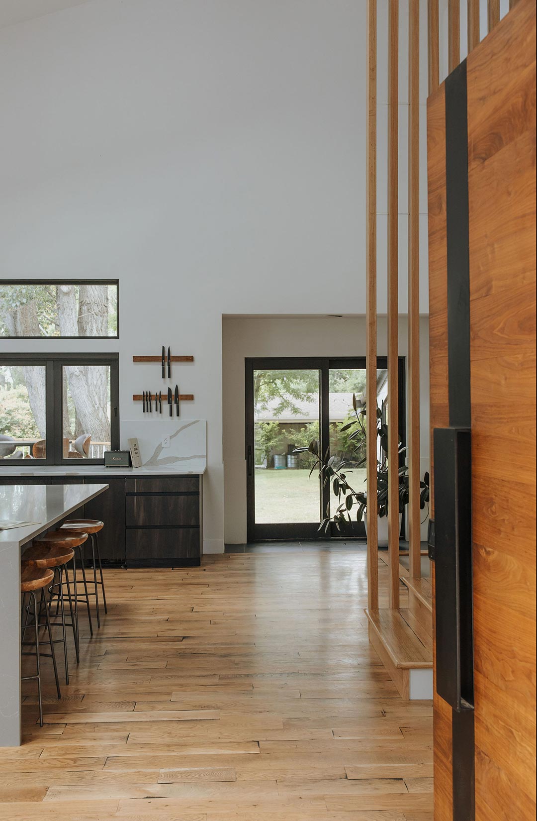 The entryway view into a rural Mid-century home designed by J. Reiko Design + Co shows a tiny bit of the horizontal wood slat entryway door. Inside you find a modern wood slat divider between the lower and upper stairs to the right and to the left a waterfall slab island with wood and metal stools.