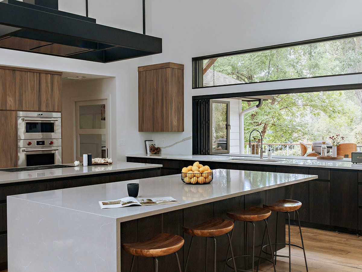 A wide-angle view of the kitchen in a rural mid-century modern remodel designed by J. Reiko Deisgn + Co. Front and center is a double floating island with countertops made from Grey Savoie Polished Quartz with a waterfall edge. The expansive accordion window that runs along the side bar cabinets is pulled open to reveal an indoor-outdoor view of a gorgeous backyard. 