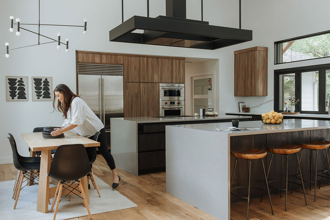 JJ. Reiko Design + Co. - Selecting Finishes - 2 Jenny Murphy, the principal designer behind J. Reiko Design + Co. places some black table wear down on a white oak, modern table in a warm toned modern kitchen when the selected finishes of waterfall concrete countertops in contrast with walnut cabinetry and modern light fixtures and a unique hood range bring the look together.