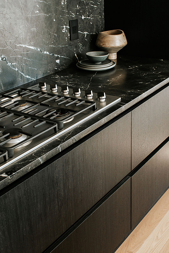 J. Reiko Design + Co. - Selecting Finishes - 6 A modern Japandi kitchen's cabinets and range are shown with earthenware on the black marble countertops with black melamine cabinets with flat surface and a chosen look of no kitchen cabinet hard wear. Japandi design by J. Reiko Design + Co.