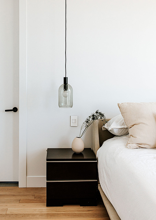 J. Reiko Design + Co. - Japandi Bedrooms - 13 Minimal Japandi bedside vignette with dark wood nightstand, glass pendant light, ceramic vase with dried stems, and layered neutral bedding.