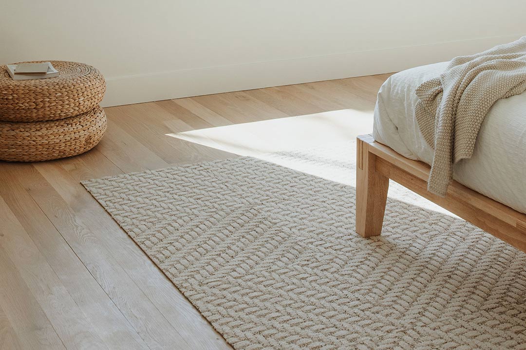 J. Reiko Design + Co. - Japandi Bedrooms - 06 Textured neutral Japandi bedroom rug beneath a natural wood platform bed with knit throw blanket and woven pouf in soft sunlight