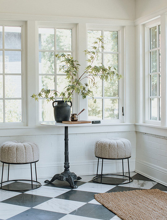 Sunroom seating vignette with café table, boucle stools, and checkerboard marble flooring.