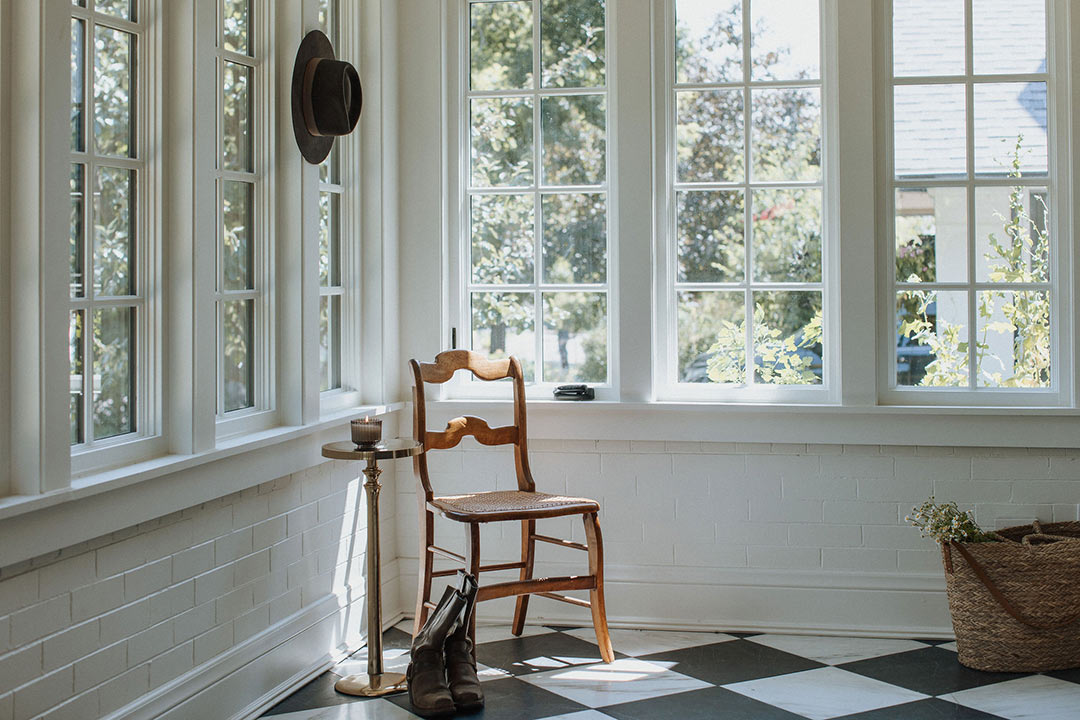 J. Reiko Design + Co. - Modern Colonial Interior Design - 31 Sunroom corner with vintage chair, boots, woven basket, and checkerboard floor by bright windows.