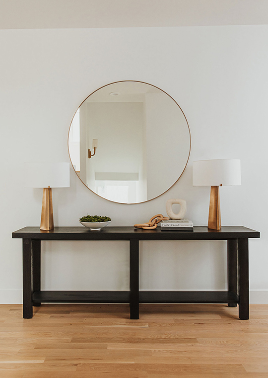 J. Reiko Design + Co. - Quiet Luxury Living Room Ideas - 12 A dark wood accent table with minimalist styling, two brass table lamps and a large circular brass mirror.