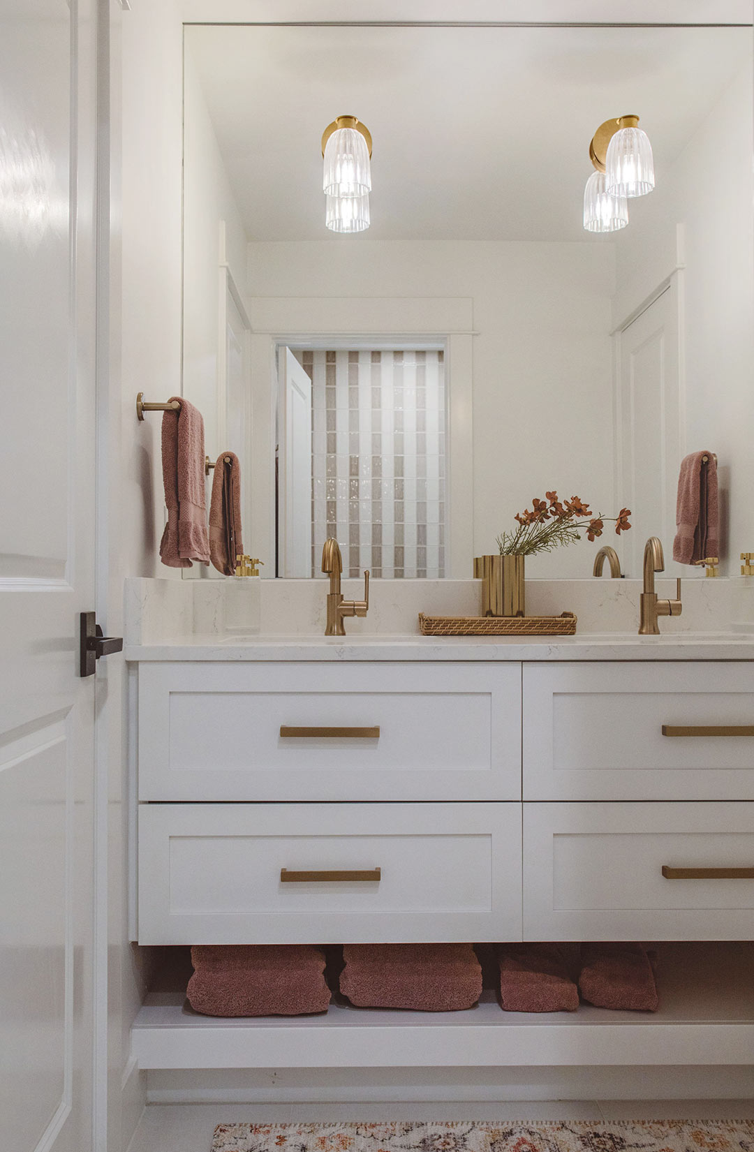 Bright bathroom with double white vanity, brass faucets, marble countertop, pink towels, and glass wall sconces above a large mirror.