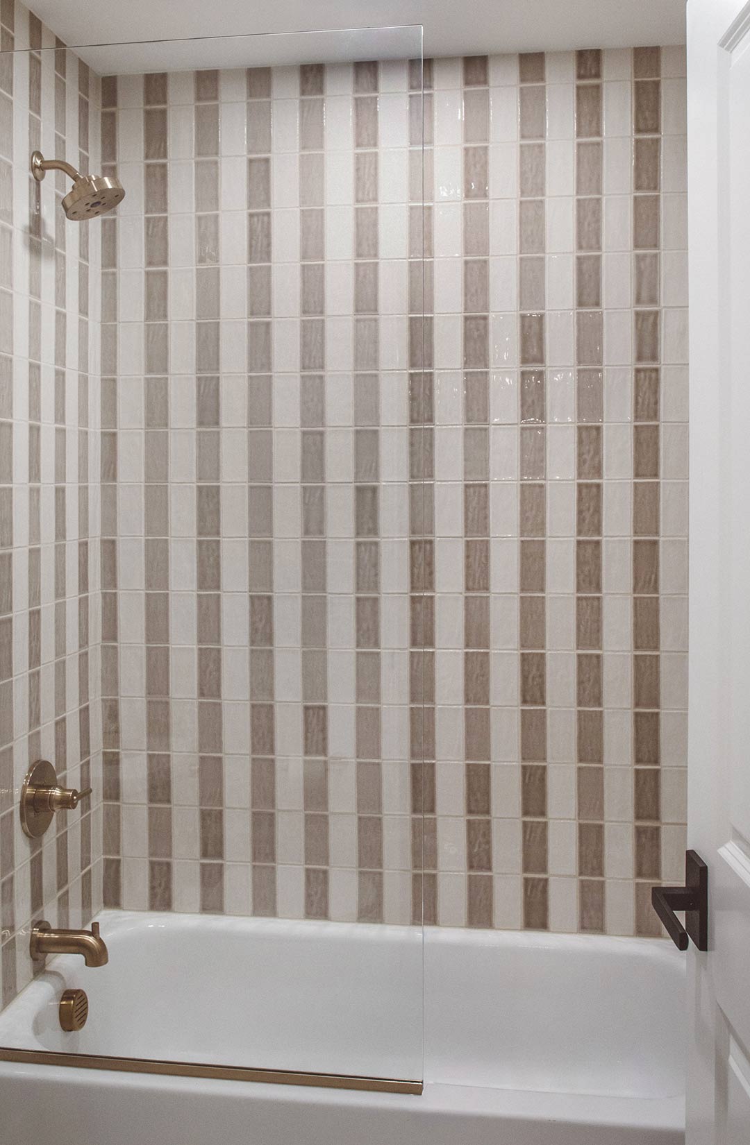 Bathtub and shower with vertical striped tile wall, glass shower panel, brass plumbing fixtures, and white tub surround in a modern bathroom design.