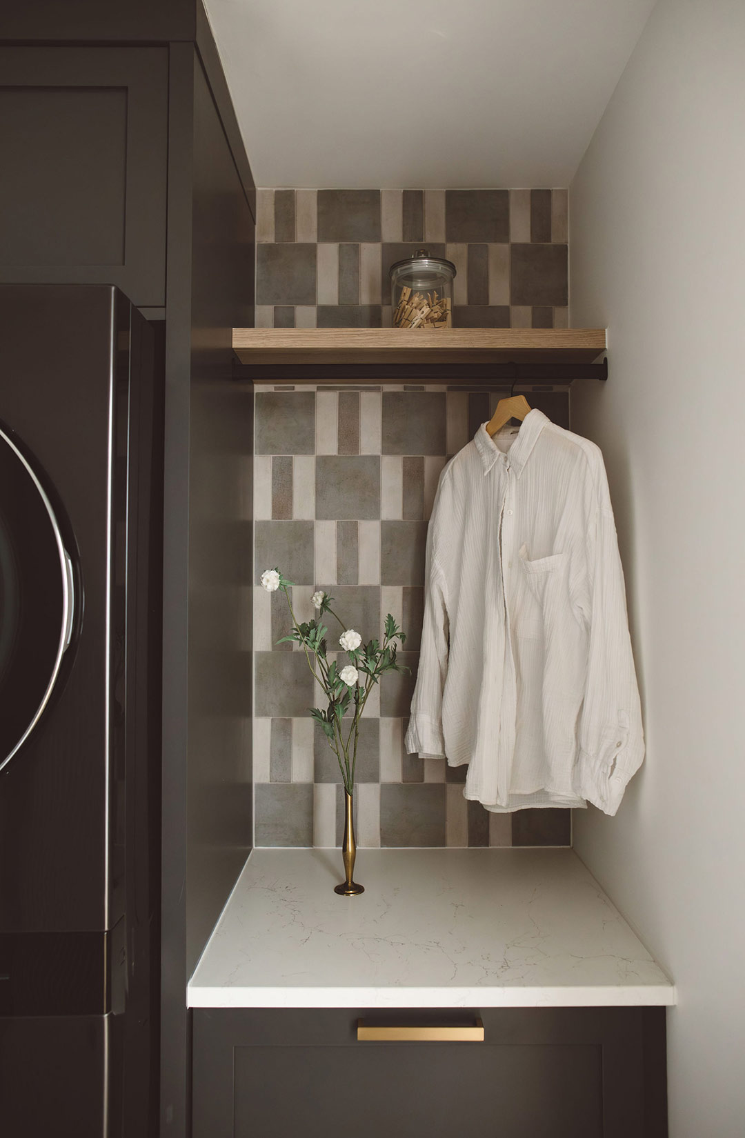 Built-in laundry nook with dark cabinetry, patterned tile backsplash, quartz countertop folding station, and wood shelf with hanging rod for laundry storage.