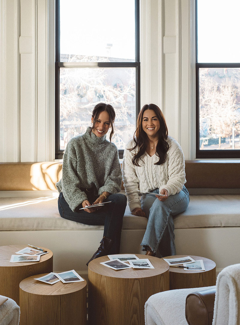 Interior designer, Jenny Murphy of J. Reiko Design + Co. sits with painter and collaborator, Natalia McGuffee in the J. Reiko Design + Co office in downtown Fort Collins. The original paintings of their collaboration spread out on the natural wood coffee table.